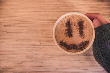 Mug of cappuccino with cinnamon for breakfast.Cinnamon powder in form of a smiley face on coffee foam.Morning cup coffee with hot cappuccino in hand.Hand in a sweater holding mug of coffee with foam.