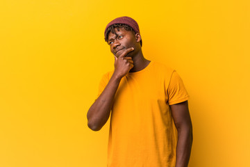 Young black man wearing rastas over yellow background looking sideways with doubtful and skeptical expression.