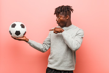 Young fitness black man holding a soccer ball impressed holding copy space on palm.