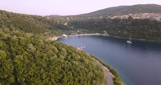 Drone shot of the coastline of greece flying towards a harbour with boats and a distant town on top of the hill surrounded by trees and forests