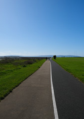 Silhouetted fighures walking down a bicycle & footpath to Salthill from Galway on a sunny summer day, with view across the bay, with blue sky.