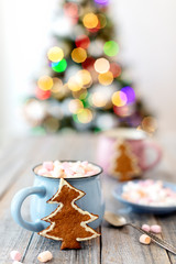 Merry Christmas! Christmas tree-shaped gingerbread cookie near cups of cocoa and marshmallows. Beautiful blurred christmas tree background with luminous garland.