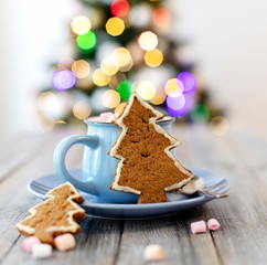 Merry Christmas! Christmas tree-shaped gingerbread cookie near cups of cocoa and marshmallows. Beautiful blurred christmas tree background with luminous garland.