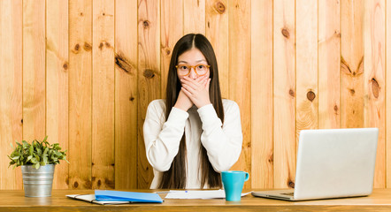 Young chinese woman studying on her desk shocked covering mouth with hands.