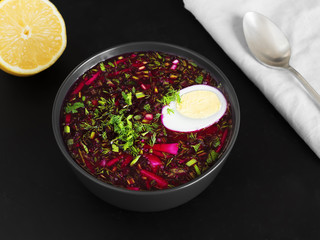Cold beetroot soup in a black ceramic plate with half a boiled egg. Next to a gray napkin is a spoon and half a lemon. Black background. Healthy, natural food. Top view.