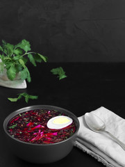 Cold beetroot soup in a black ceramic plate with half a boiled egg. A bowl on a gray napkin, next to a spoon and sprigs of cilantro. Copy space. Black background. Healthy, natural food. Close-up.