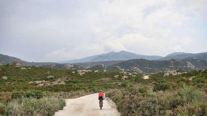 Obraz premium Cycling. Offroad Cycling in Agriates Desert, Corsica, France. a red shirt cyclist, Dusty road, Rocky road, Mediterranean Vegetation. Mountains, Daylight. Cloudy sky. Courage for the Journey. 