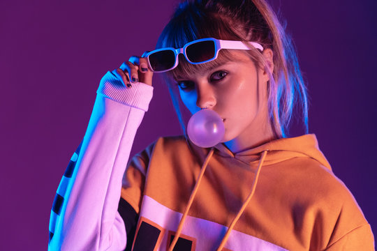 Stylish pretty young 20s fashion teen girl model wear glasses blowing bubble gum looking at camera standing at purple studio background, igen teenager in trendy night glow 80s 90s concept, portrait