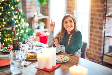 Young beautiful woman sitting eating food around christmas tree at home with a big smile on face, pointing with hand and finger to the side looking at the camera.