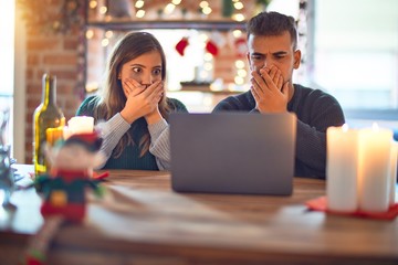 Young beautiful couple sitting using laptop around christmas decoration at home shocked covering mouth with hands for mistake. Secret concept.