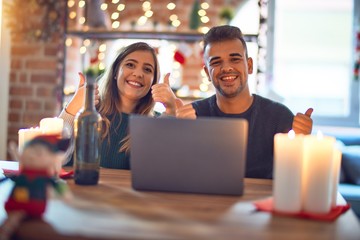 Young beautiful couple sitting using laptop around christmas decoration at home success sign doing positive gesture with hand, thumbs up smiling and happy. Cheerful expression and winner gesture.
