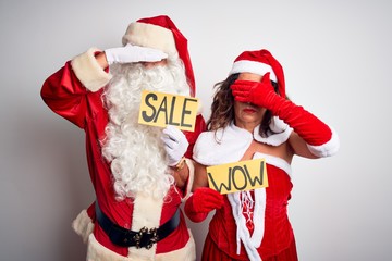 Couple wearing Santa costume holding wow and sale banner over isolated white background covering eyes with hand, looking serious and sad. Sightless, hiding and rejection concept
