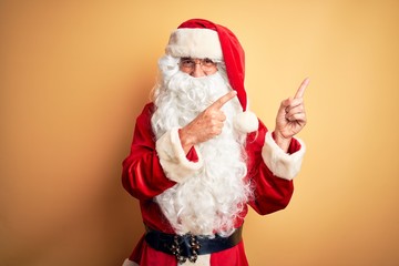 Middle age handsome man wearing Santa costume standing over isolated yellow background smiling and looking at the camera pointing with two hands and fingers to the side.