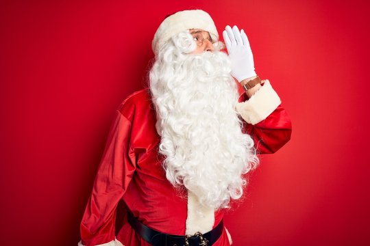 Middle age handsome man wearing Santa costume standing over isolated red background shouting and screaming loud to side with hand on mouth. Communication concept.