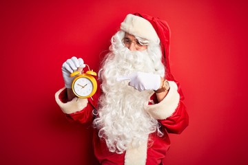 Senior man wearing Santa Claus costume holding alarm clock over isolated red background very happy pointing with hand and finger