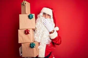 Senior man wearing Santa Claus costume holding tower of gifts over isolated red background cover mouth with hand shocked with shame for mistake, expression of fear, scared in silence, secret concept