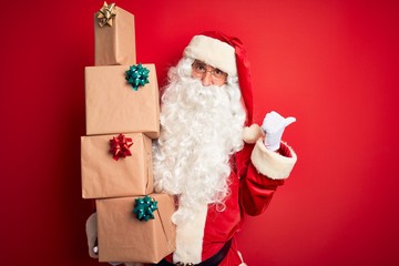 Obraz na płótnie Canvas Senior man wearing Santa Claus costume holding tower of gifts over isolated red background pointing and showing with thumb up to the side with happy face smiling