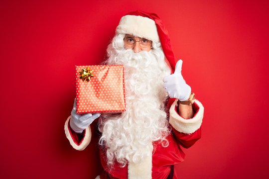 Middle age man wearing Santa Claus costume holding gift over isolated red background happy with big smile doing ok sign, thumb up with fingers, excellent sign