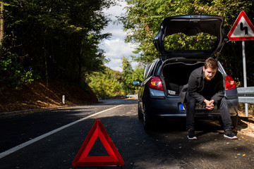 A broken car on the road, the driver holds an open hood and watches what has broken down, a roadside assistance is waiting