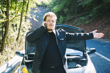 A broken car on the road, the driver holds an open hood and watches what has broken down, a roadside assistance is waiting