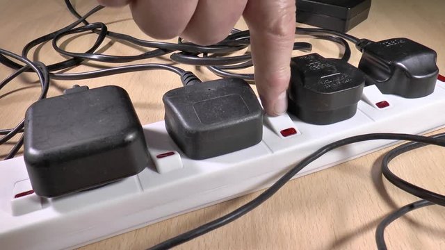 Close POV shot of a man’s hand switching on four plugs in a UK plug board, with their trailing wires lying untidily around.