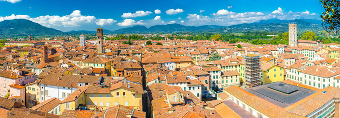 Aerial top panoramic view of historical centre medieval town Lucca with old buildings, typical orange terracotta tiled roofs and mountain range, hills, blue sky white clouds background, Tuscany, Italy - obrazy, fototapety, plakaty