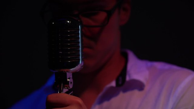 Young Caucasian man in a white open shirt with short hair in glasses lowers, regulate and fix the classic retro glare microphone on stage in dark empty club close up. Male walks away. Spotlights shine