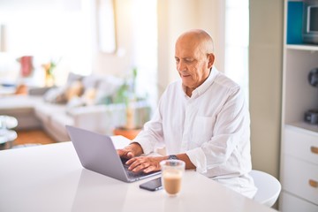 Senior handsome man smiling happy and confident. Sitting using laptop and drinking cup of coffee at home