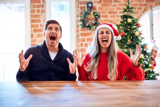 Young couple wearing santa claus hat sitting on chair and table around christmas tree at home crazy and mad shouting and yelling with aggressive expression and arms raised. Frustration concept.