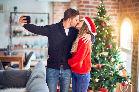 Young beautiful couple smiling happy and confident. Hugging and kissing make selfie by camera standing around christmas tree at home