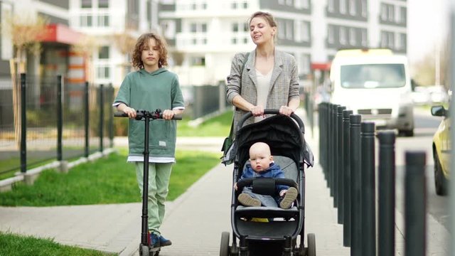 Beautiful young woman with a stroller walking with his sons on the city street. Kinky schoolboy rides an electric scooter. Mothers Day