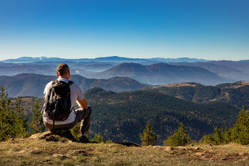 Rear view of male hiker with backpack sitting on top of the mountain and enjoying the view during the day.