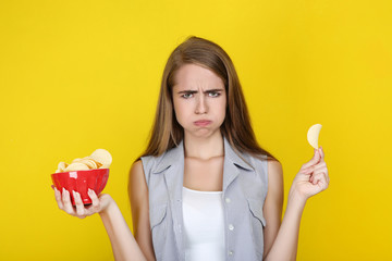 Sad girl with potato chips in bowl on yellow background