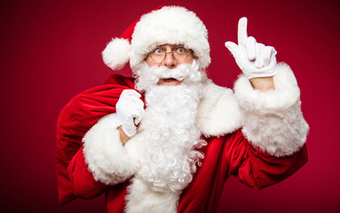 I have an idea! Close up photo of Santa, posing with a surprised face expression, holding a sack of presents in his right hand and pointing up with his left index finger.