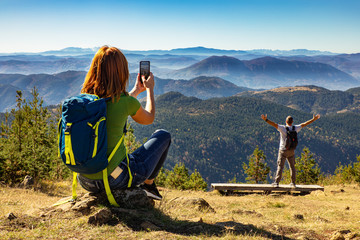 Hikers couple with backpack on top of the mountain, woman photographing her husband with phone.