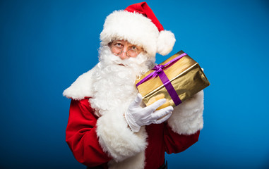 Look at your gift! Authentic Santa Claus is showing a box in a golden wrapping paper with a purple ribbon on it, looking at the camera happily.
