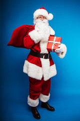 Happy Santa. Full-length photo of Santa Claus, posing with a sack of presents on his right shoulder and a present in a red wrapping paper in his right arm, looking at the camera.