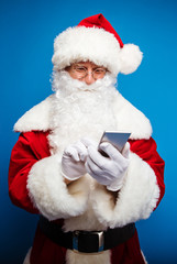 Checking my list. Authentic Santa Claus is holding a smartphone in his right hand, reading something from the screen and tapping on it.