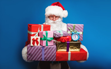 Timeless kindness. Authentic Santa is posing with a pile of multicolored Christmas gifts and a sky blue alarm clock  in his hands, looking at the camera over his round-shaped glasses.