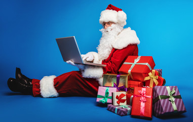 Buying presents online. Authentic Santa Claus is sitting on the floor near a pile of boxes in multicolored wrapping paper while typing something on his laptop.