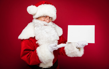 A Christmas postcard. Santa Claus pointing with his right hand at the blank postcard with copyspace on it, which he holds in his left hand, while looking at the camera.