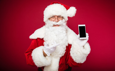 Your present. Happy Santa Claus is posing, showing a smartphone in his left hand, and pointing at it with his right index finger.