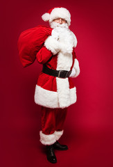 Father Christmas. A full-length photo of Santa Claus, posing on a red background holding a sack full of presents on his right shoulder and his left hand on the belt.