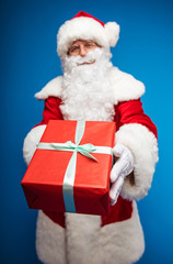 A gift. Photo of Santa Claus, looking at the camera and holding a red present box with a turquoise ribbon in his outstretched arms.