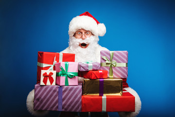 Gifts for all family. Real Santa Claus is holding a pile of presents of various colors in his hands, looking at the camera with surprised face expression.