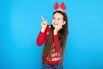 Little girl in christmas sweater and red horns pointing by finger on blue background