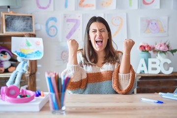 Young beautiful teacher woman wearing sweater and glasses sitting on desk at kindergarten very happy and excited doing winner gesture with arms raised, smiling and screaming for success. 