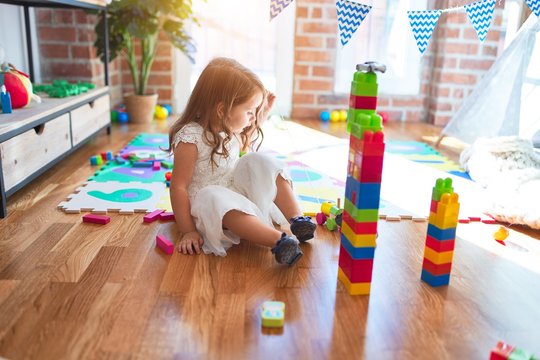 Adorable blonde toddler playing with building blocks around lots of toys at kindergarten