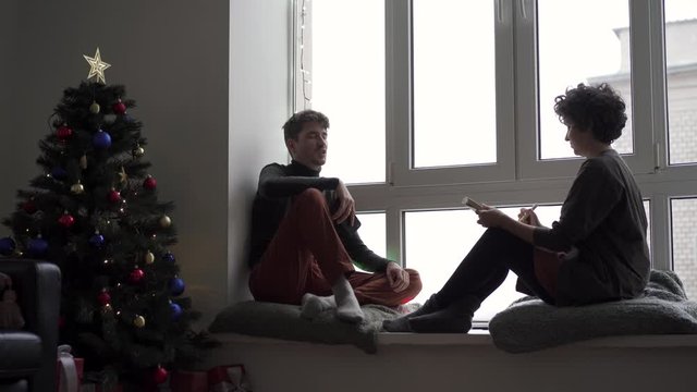 A young couple sits talking and chatting at a large window next to a Christmas tree in their cozy home. A girl draws a man or makes sketches.