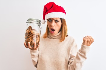 Beautiful redhead woman wearing christmas hat holding cookies jar over isolated background annoyed and frustrated shouting with anger, crazy and yelling with raised hand, anger concept